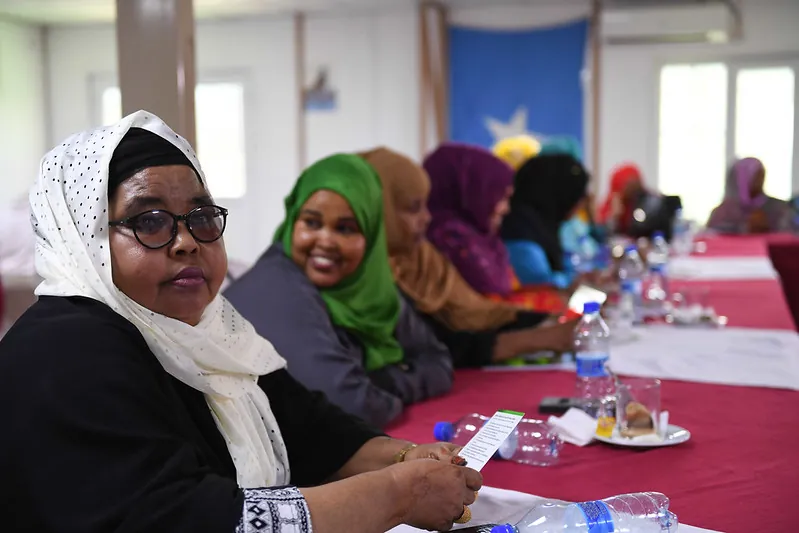 Lawmakers wearing head scarves sit at a long conference table set with bottles of water and small plates of food.