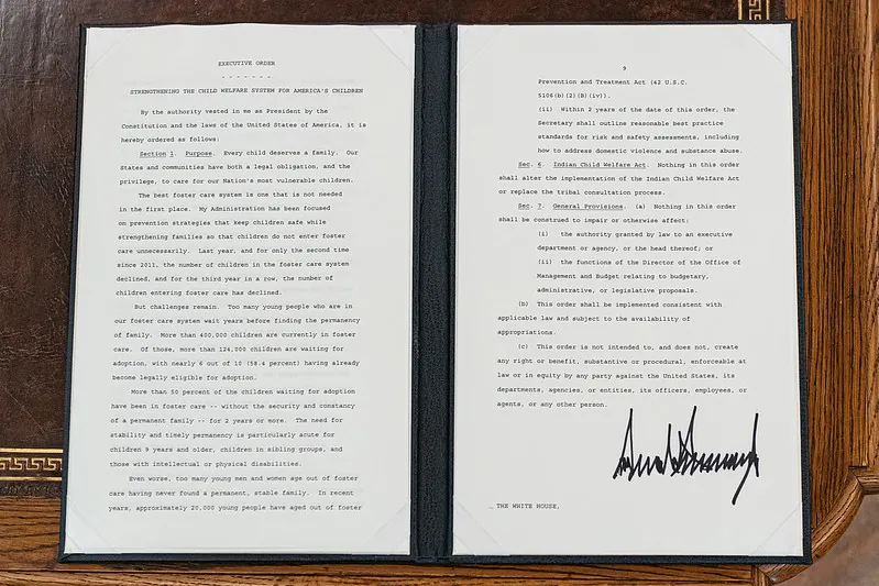 An open portfolio on a desk shows side-by-side sheets of paper typed with an executive order issued by the President of the United States and signed in the lower right by then-president Donald Trump.