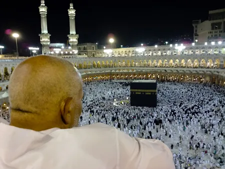 A man dressed in white is shown from behind looking down over the Kaaba, Islam’s most sacred site. Hundreds of other people, dressed in all black or all white, can be seen circling a large black cube-like structure on the floor of a stadium-like structure. 