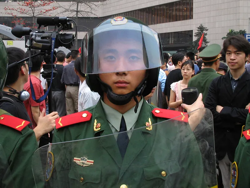 A uniformed police officer from the Chinese People’s Armed Police Force wears a helmet with a face shield and holds a clear plastic shield. The officer is surrounded by people, including police officers, news media, and other people standing outside a building.