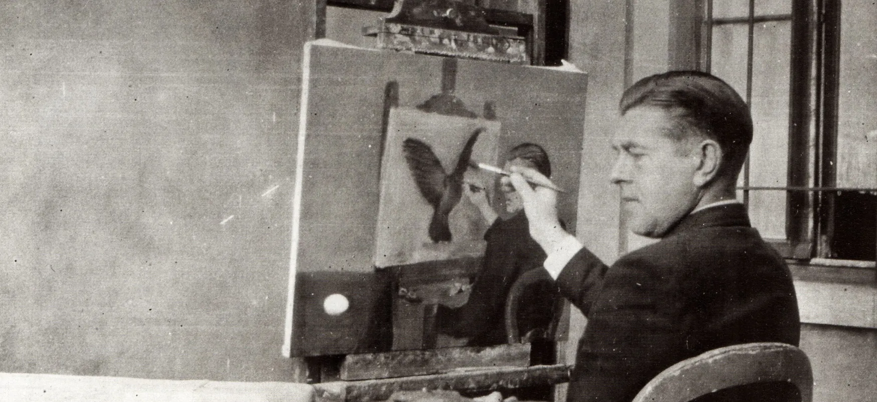 Black and white photograph of a seated man holding a paint brush to a nearly finished canvas resting on an easel.