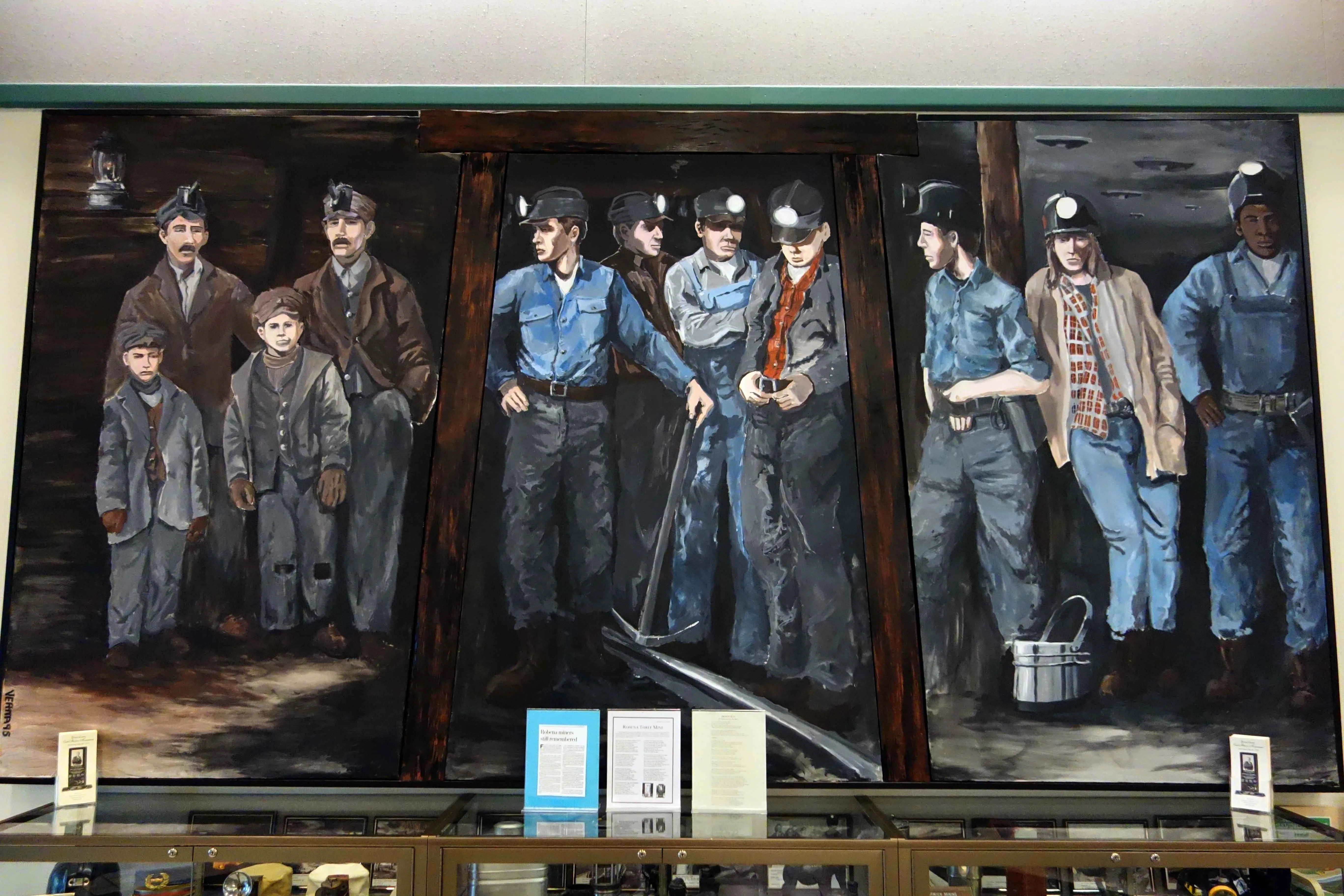 A painting depicts three eras of coal mining. The first appears to be 100 plus years ago, and children are in the mine alongside men. In the era are men with headlamps and pitchforks. In what appears to be the most recent era there are men standing in a mine that appears to be more sturdily built.