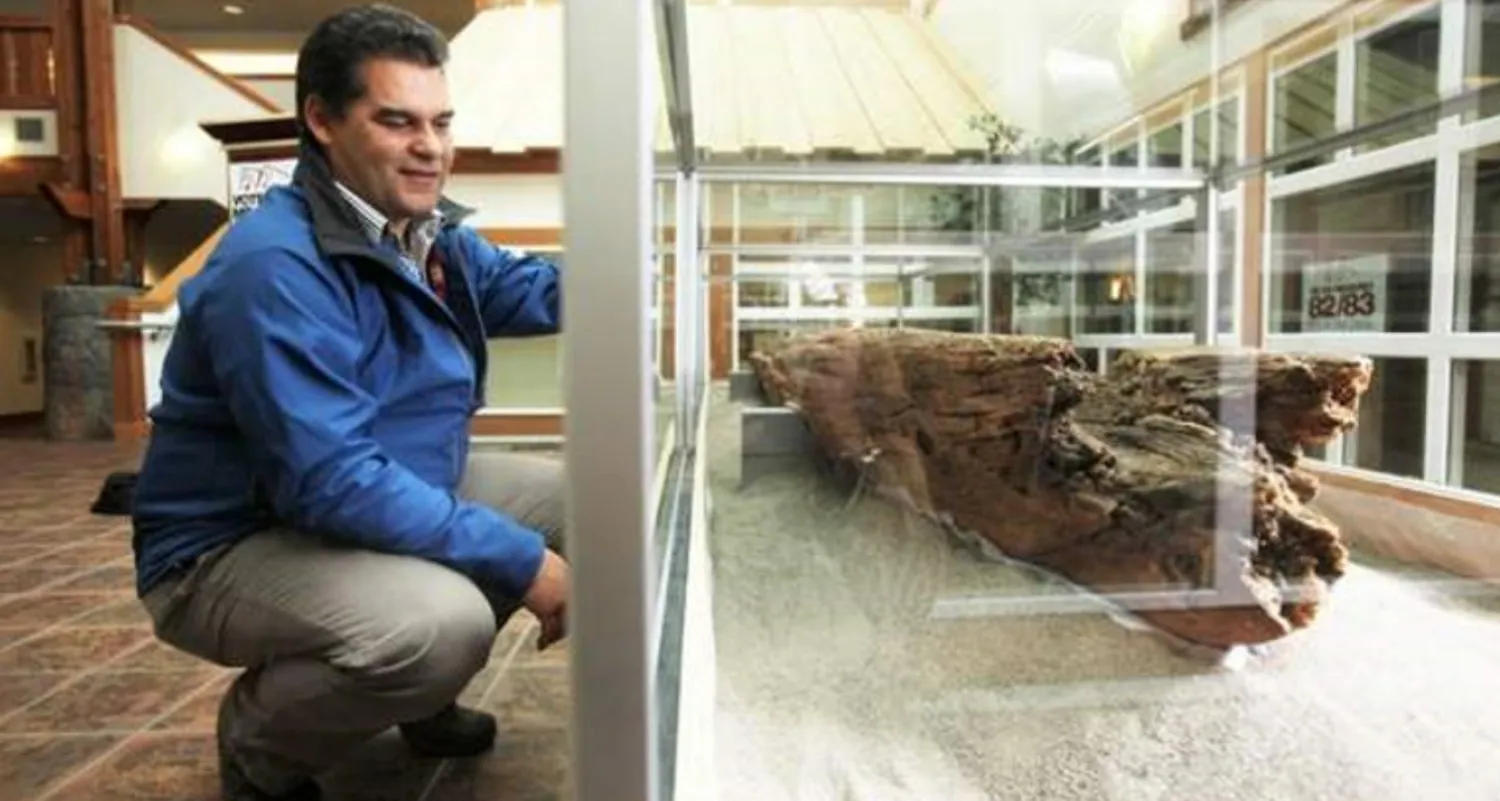 A man squatting down and looking at an old canoe in a glass display case.