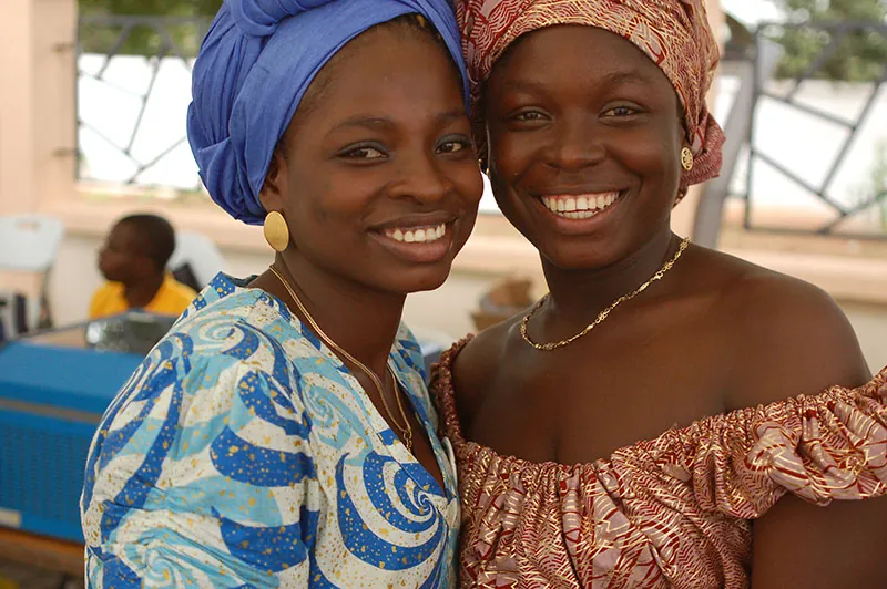 Two Ghanaian women are standing with their heads touching facing forward. Both are smiling. The woman on the left is wearing a blue head scarf and a blue patterned dress. The woman on the right is wearing an orange patterned head scarf and an off the shoulder dress of the same pattern. Both women are wearing simple gold chain necklaces and earrings.