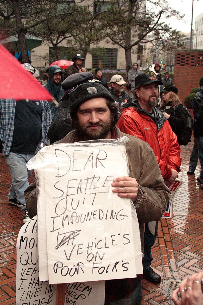 A photo shows a man protester at an anti-war protest rally, holding a placard that reads, “Dear Seattle Quit Impounding Vehicles on Poor Folks.”