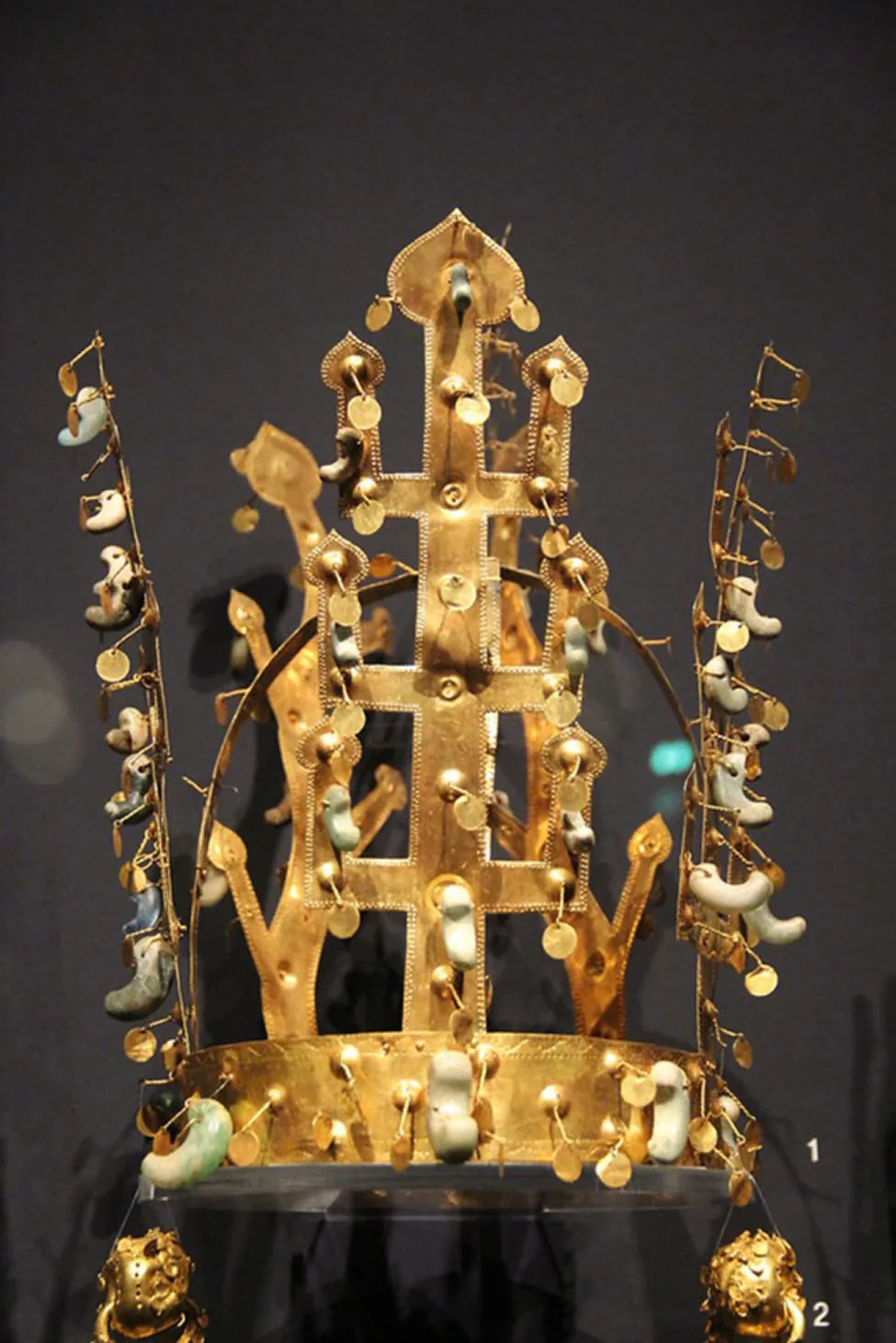 A picture of a gold crown. The crown has etchings along the edges throughout. Two tall pieces stick up at each side of the bottom round band and in the middle of the crown a tall piece sticks upright with three elbow projections on each side with leaf-like decor at the ends as well as at the top. The back of the crown has two long stems sticking upright with four similar leaf-like endings. A piece goes from the left and bends and connects on the right side. Small knobs are seen throughout the crown with small “C” shaped white and pale blue and green objects in varying sizes attached with gold or white strings.