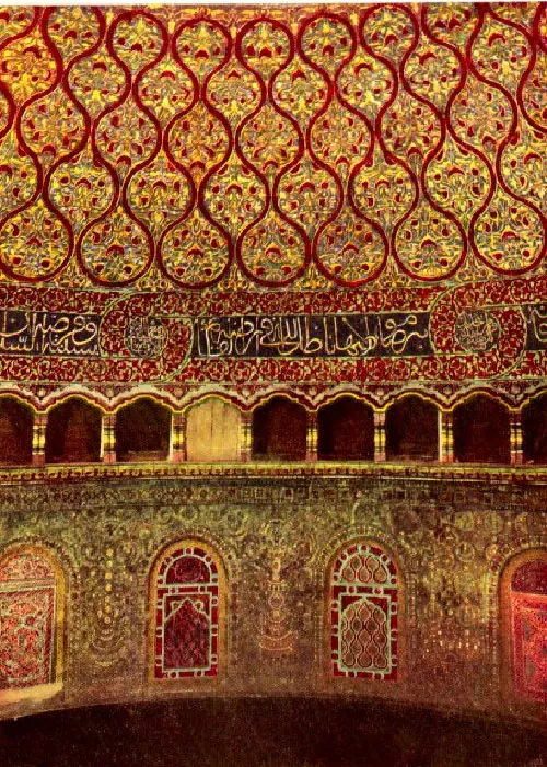 A picture of a rounded, red and gold mosaic wall in shown. The top half of the wall shows barbell shaped red lines surrounding red and gold mosaic tiles. A thick red band of tile runs below with an inscription in scripted writing running in the middle in gold on black tile. Richly decorated arched openings show below that in gold and red and at the bottom a highly decorated wall is seen with colorful tiles and arched sections with more decorated tiles.