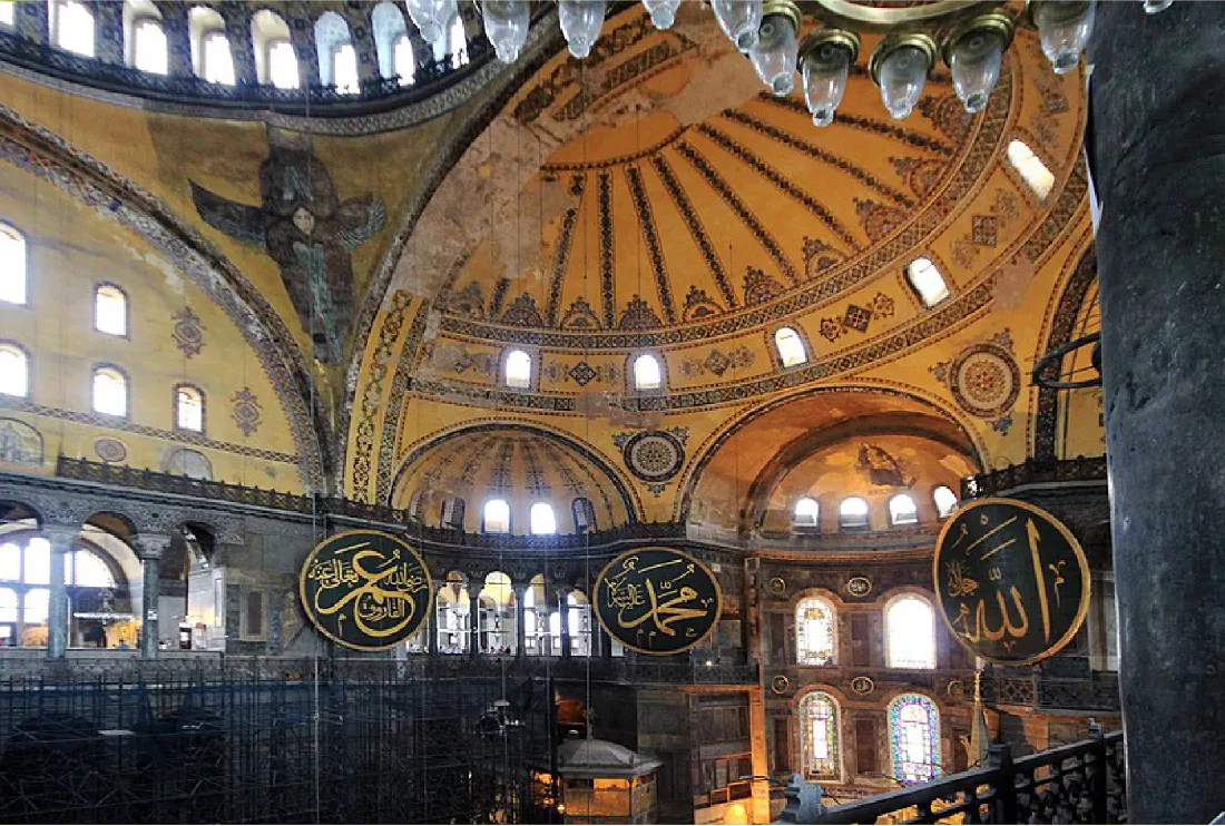 This photograph shows the interior of Hagia Sophia. The building has richly decorated dome ceilings. Medallions bearing Arabic writing are on the walls.