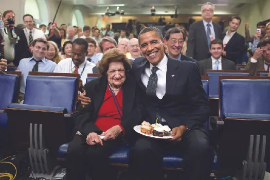 An image of Barack Obama and Helen Thomas seated. Obama holds a plate of cake.