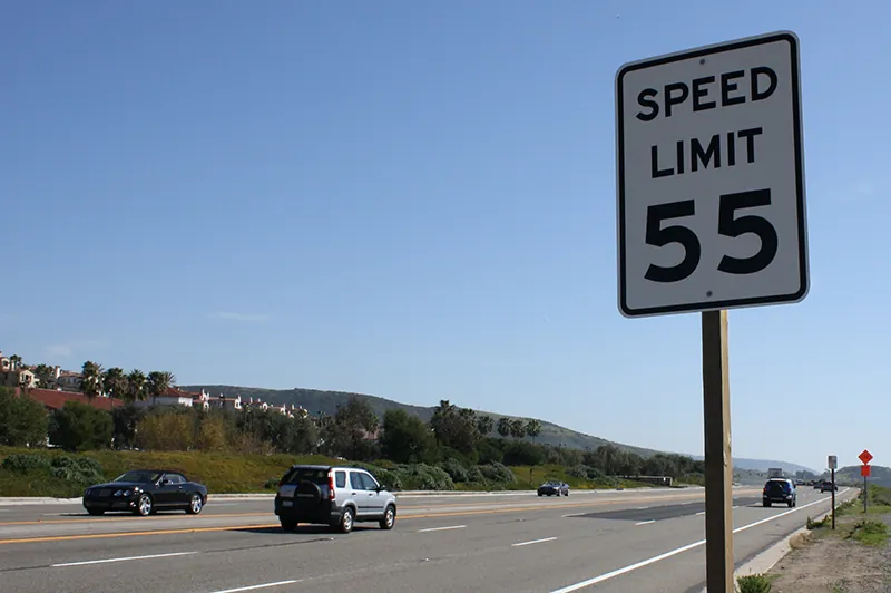 A sign on the side of a road indicates the speed limit is 55 miles per hour. On the road, cars travel in both directions.