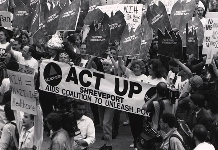 A large street demonstration protests the National Institutes of Health (NIH). Several protestors hold a banner that reads “ACT UP Shreveport AIDS Coalition to Unleash Power.”