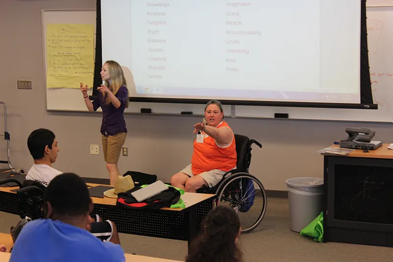 A photo shows a woman lecturing disabled students in a lecture hall, with a large projector screen in the background. Another woman in wheelchair appears by her side trying to interact with the students.