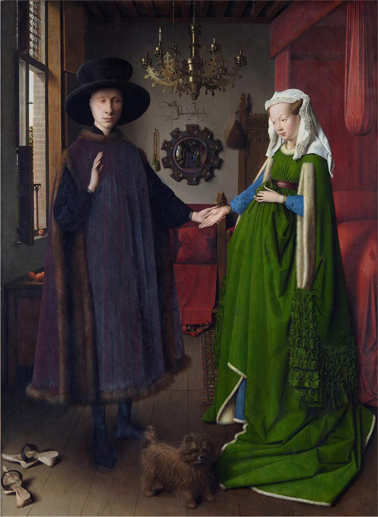 An image of a painting is shown. A man and a woman are shown standing in a room with a dark red bed with a canopy on the right and a red covered wood piece of furniture in the background. The floor is brown wood and a richly decorated rug can be seen at the foot of the bed. A large round mirror hangs on the back wall with notches in the dark frame. A chandelier hangs down from the ceiling in the middle and a window is seen at the left. Round orange objects are seen laying on the window sill as well as on a table in front of the window. The man is on the left and wears a large black brimmed hat, has a pale face, large nose, and wears long blue and purple robes trimmed in fur at the edges. His left hand extends to the woman whose right hand is palm facing up in his left hand. She wears her brown hair up in cones at the sides of her head covered in a lace trimmed white cloth, long rich green layered dress with blue sleeves showing out from the cloak. She has a pale face and wears a necklace. A small brown furry animal with a short tail is at their feet and a pair of clogs with black straps is shown in the bottom left corner of the image.