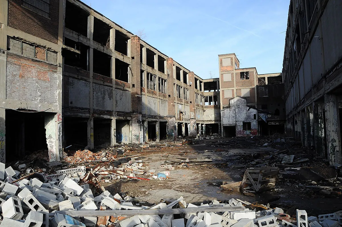 A large open space within a factory complex is littered with discarded cinder blocks, garbage, and pieces that have broken off the buildings. The structures all have broken windows and eroding concrete.