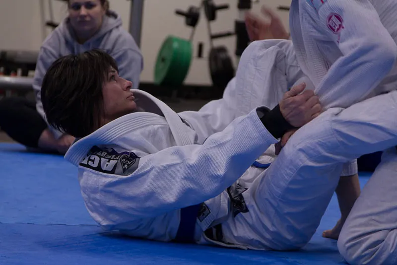 A photo shows two persons in jiu-jitsu uniforms sparring during a jiu-jitsu training session as another woman in the background watches them.