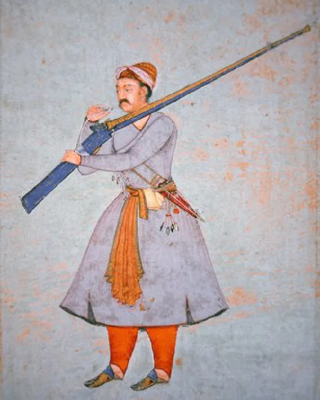A man with a black moustache wears a long, blue belted shirt with two slits along the bottom, red pants, and ornate blue and orange slippers. On his head he wears an orange hat with a white cloth tied around it. He holds a blue and orange rifle over his left shoulder with his left arm and is putting something on a string toward his mouth with his right. His belt holds many colorful, hanging objects as well as a long orange scarf.
