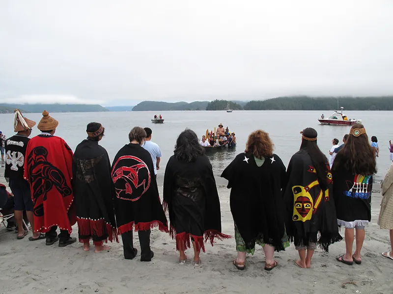 A group of people stand at the edge of a lake wearing long ponchos woven with First Nations symbols. They watch as A-in-chut (Shawn Atleo) returns to shore in a canoe.