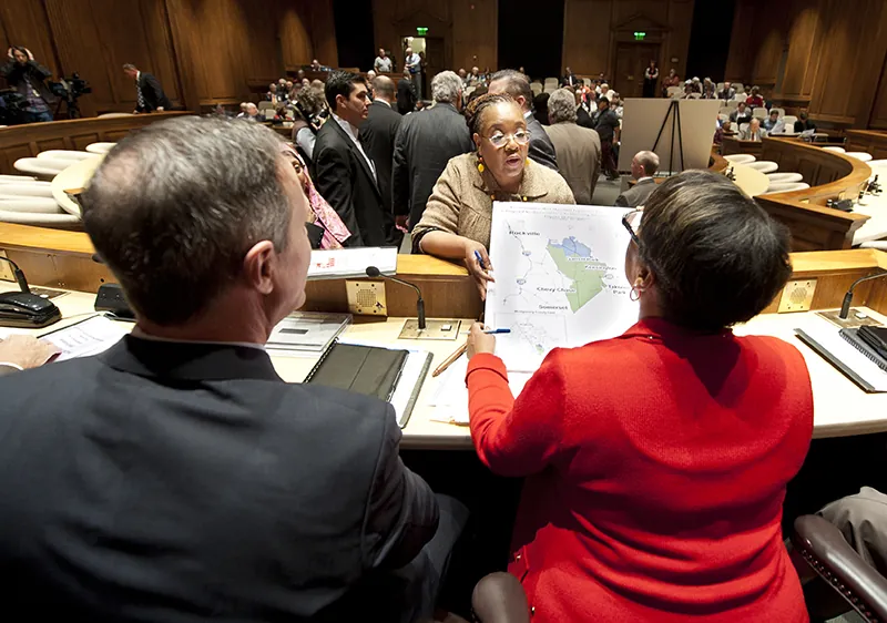 Two people wearing suits sit behind a dais and look at a document showing a map that  another person hands to them from the other side of the dais.  Other people stand around the room talking.