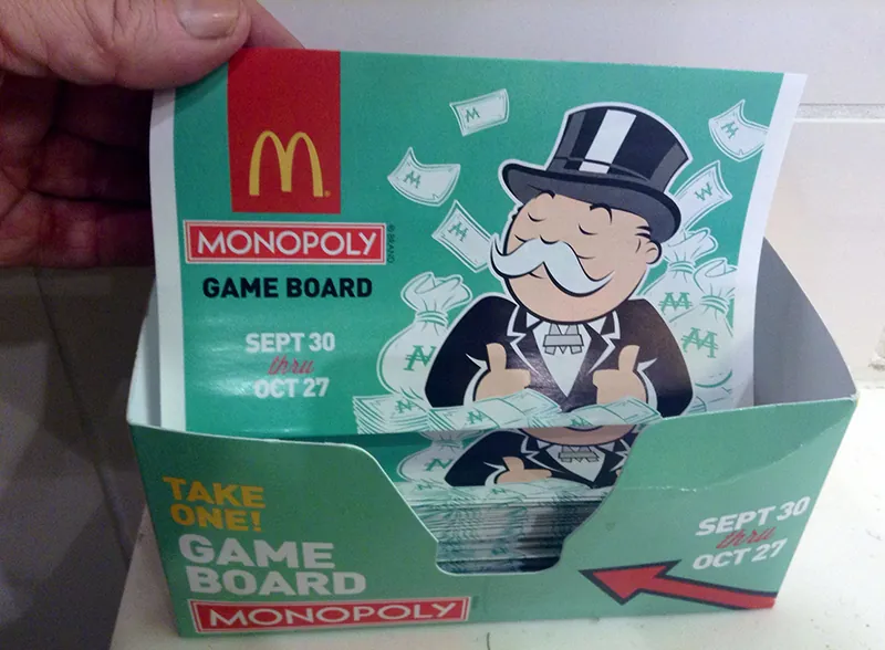 A small cardboard display box contains McDonalds Monopoly gameboards. The box says “Take one! Game Board Monopoly. September 30 through October 27”. An arrow points to the gameboards. Similar copy is written on the folded up gameboard, along with a picture of the Monopoly Man in his top hat and suit.