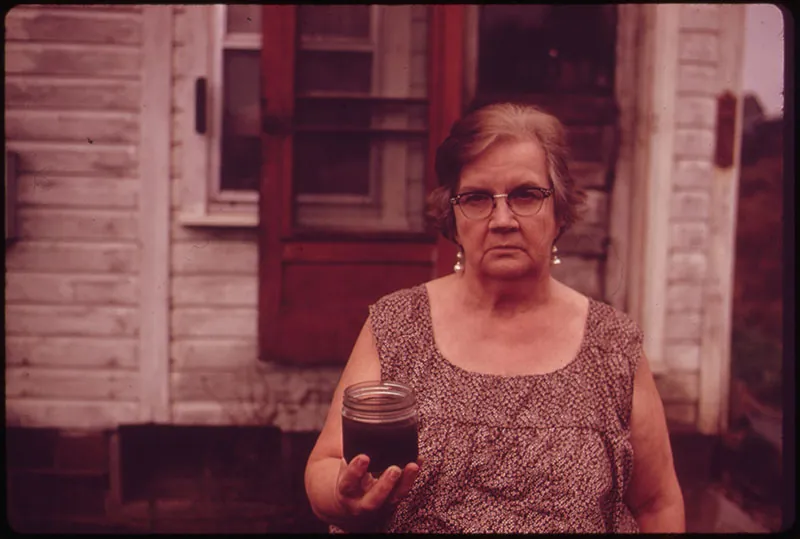 Standing in front of a dilapidated home, a citizen holds a jar filled with brown water.