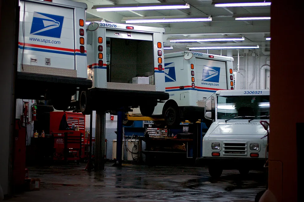 Se muestra una foto de un taller mecánico de automóviles. Hay tres camiones del Servicio Postal de Estados Unidos que están en mantenimiento y uno que no lo está.