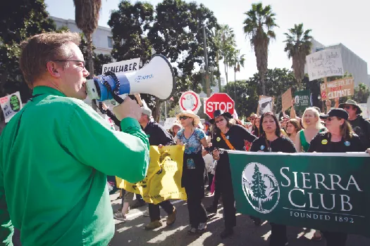 An image of a person speaking through a bullhorn on the left, and a crowd of people marching down a street on the right. Several marchers are holding a large banner that reads “Sierra Club”.