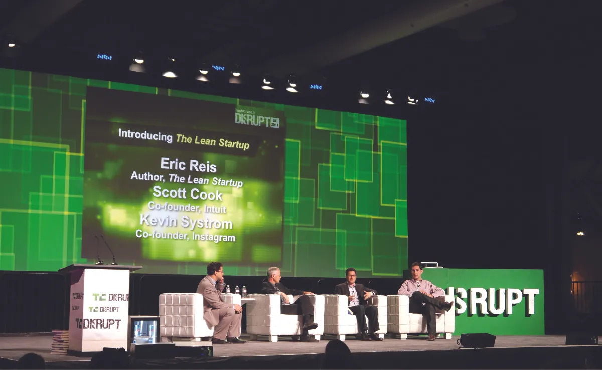 Four men sitting on a stage talking about lean startup. A screen shows the names Eric Ries (author, The Lean Startup), Scott Cook (co-founder, Intuit), and Kevin Systrom (co-founder, Instagram).