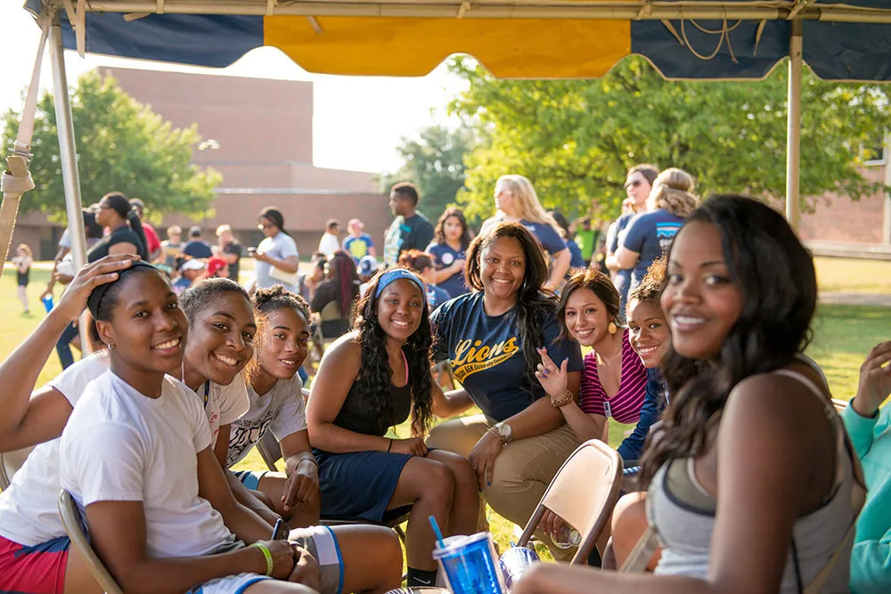 A group of young women sit outside under a tent. Behind them are other groups. 