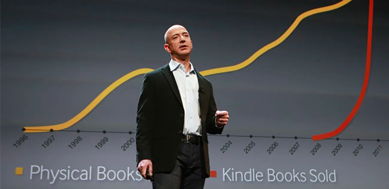 A photo shows Jeff Bezos flashing the slide showing the phenomenal growth of Amazon’s Kindle eBook sales in comparison to physical book sales during his presentation of the new Kindles.
