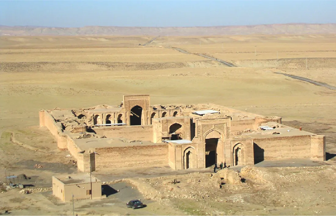 A picture of a large, open, square fortress-like structure is shown in the middle of a barren desert. The front shows a tall, open, rectangular entrance with smaller ornately carved doors to either side. Another ornate doorway in a wall is shown running down the middle of the structure. The back wall shows another tall open doorway with three smaller doorways on either side. Most of the structure is open with only the four corners showing clay roofs in irregular dimensions. Two cloaked people are shown walking into the main doorway. A small square, metal building is located in the front left of the picture and a car is shown parked in the forefront. One road is shown in the right of the picture leading into the background.