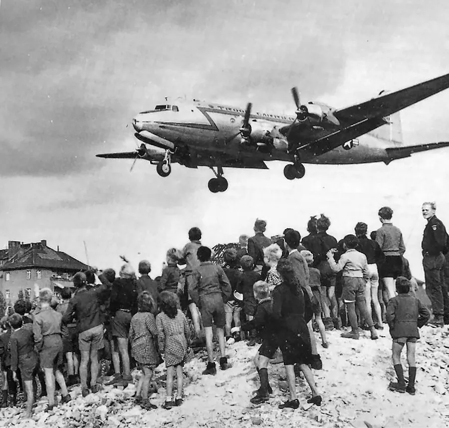 A photograph shows a cargo airplane with two propellers on each wing and its landing wheels down flying closely over a large crowd of people. Men, women, and children are standing on a hill as the plane passes above them.