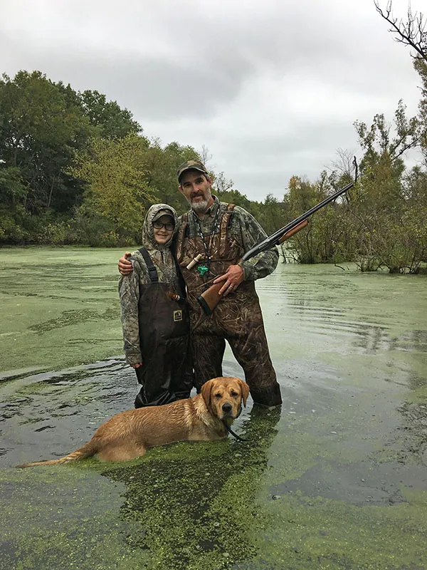 An adult and young teen stand in an algae-covered river wearing waders and camouflage; the adult is holding a shotgun.