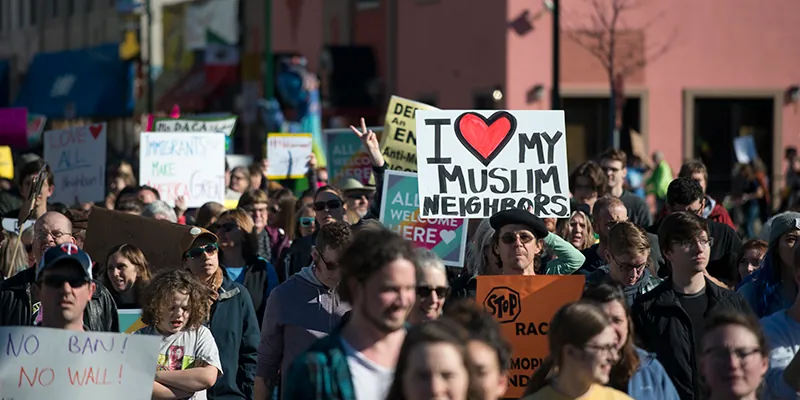 Color photograph of a crowd of people filling a city street. Many carry signs. Prominent in the center of the photograph is a large hand-written sign reading "I Love My Muslim Neighbors." Also visible is a raised hand displaying the peace symbol.