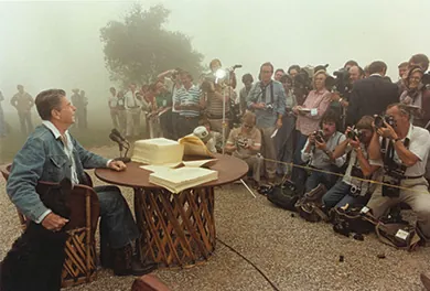 A photograph shows Ronald Reagan signing legislation while seated outdoors at a rustic table. He is dressed in blue jeans, a denim jacket, and cowboy boots, and he strokes the head of a large black dog seated beside him. In front of Reagan, the press takes photographs.