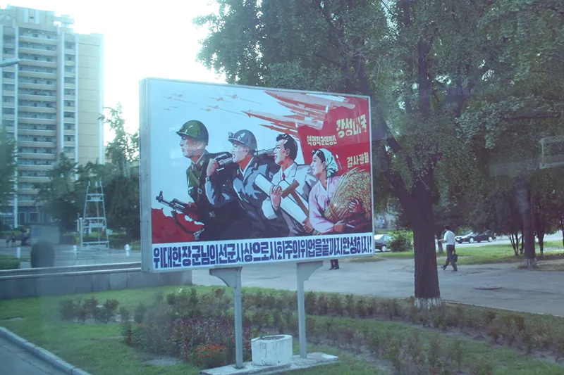 A propaganda poster in the middle of a neighborhood shows four people: a uniformed soldier wearing a helmet and carrying a gun is in the lead, followed by a worker wearing a helmet and coveralls, followed by a person in a suit carrying a roll of paper and a t-square, followed by a peasant farmer holding grains. Stylized military jets fly and missiles appear ready to deploy over their heads.