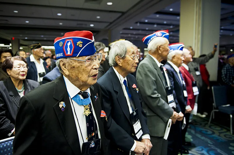 A group of decorated Japanese World War Two veterans stand in rows, wearing name tags on lanyards, in a large carpeted event space.