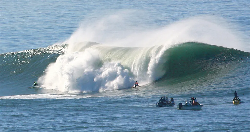 An ocean whitecap crashes near two surfers and three boats.