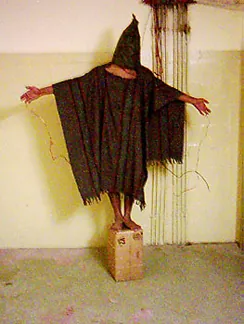 Zdjęcie przedstawia osobę stojącą na pudełku z wyciągniętymi w bok ramionami. Osoba zakryta jest czymś w rodzaju narzutki, a jej głowa schowana jest całkowicie pod kapturem.