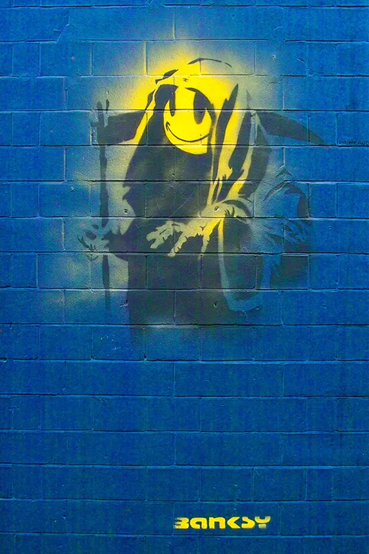 Spray painted image on brick wall of Grim Reaper figure with a bright yellow smiley face.