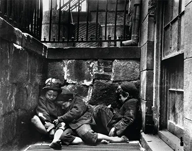 A photograph shows three small children, shabbily dressed and barefoot, asleep in a heap over a steam grate slightly below street level.