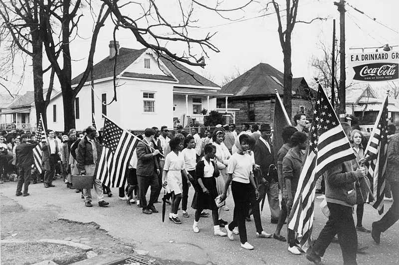 A large group of people, including many Black people as well as a few White people, march down a street. They walk 4 to 5 people across, and some carry large American flags.