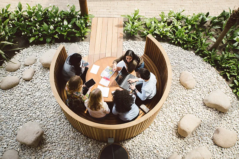 A photo shows an overhead view of students sitting in a round wooden cubicle built in the winter garden of the Essex Business School campus. The area around the cubicle is decorated with white pebbles and stones bounded by small green plants.