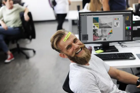 Na pierwszym planie widoczny jest szeroko uśmiechnięty brodaty mężczyzna po trzydziestce, który siedzi przy komputerze. Na czole ma naklejoną kartkę „be happy”.
