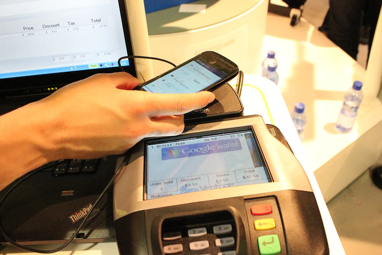 A photo shows a point of sales machine at a store. The screen on the machine reads, google wallet, and a person is holding their phone above the flat base next to the screen.