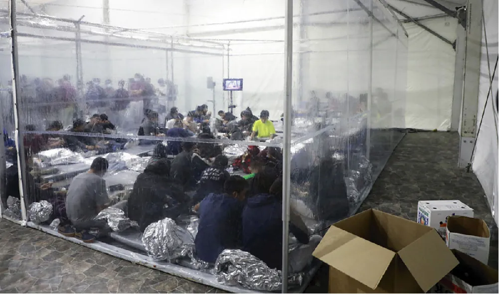 About twenty young people sit on the floor of a holding area with plastic sheeting as walls. Plastic blankets litter the floor, and boxes are outside. In the background a similar holding area with more people can be seen.