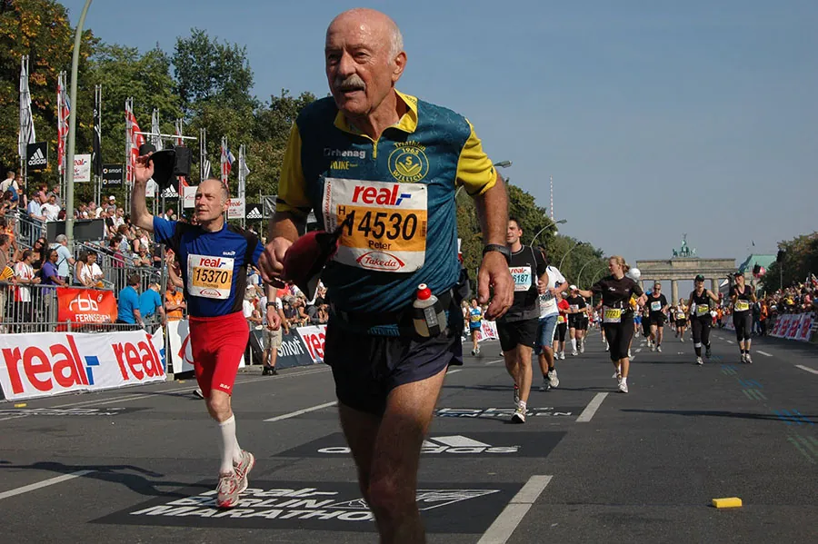 A group of people run a marathon in Berlin, Germany. The Brandenberg Gate is in the background. In the immediate foreground and ahead of many other runners, there is runner who appears much older than the others.
