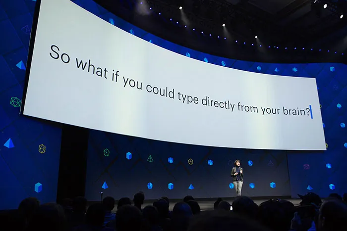 Facebook VP of Engineering Regina Dugan stands on a stage with a large screen above her. The text reads, 'So what if you could type directly from your brain.’