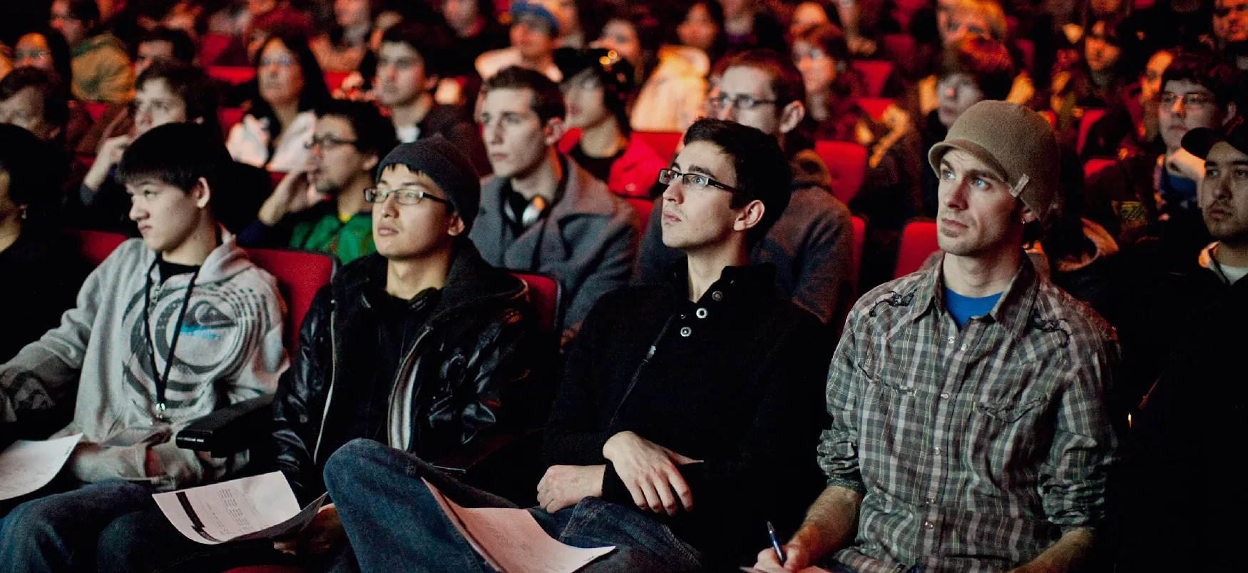 An audience sits in a theater watching movie. The four young people sitting in the first row are taking notes.
