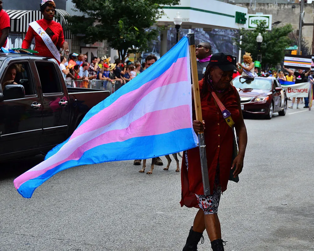 A person carries a transgender pride flag in a parade. Behind them is a person stands in the bed of a pickup truck.