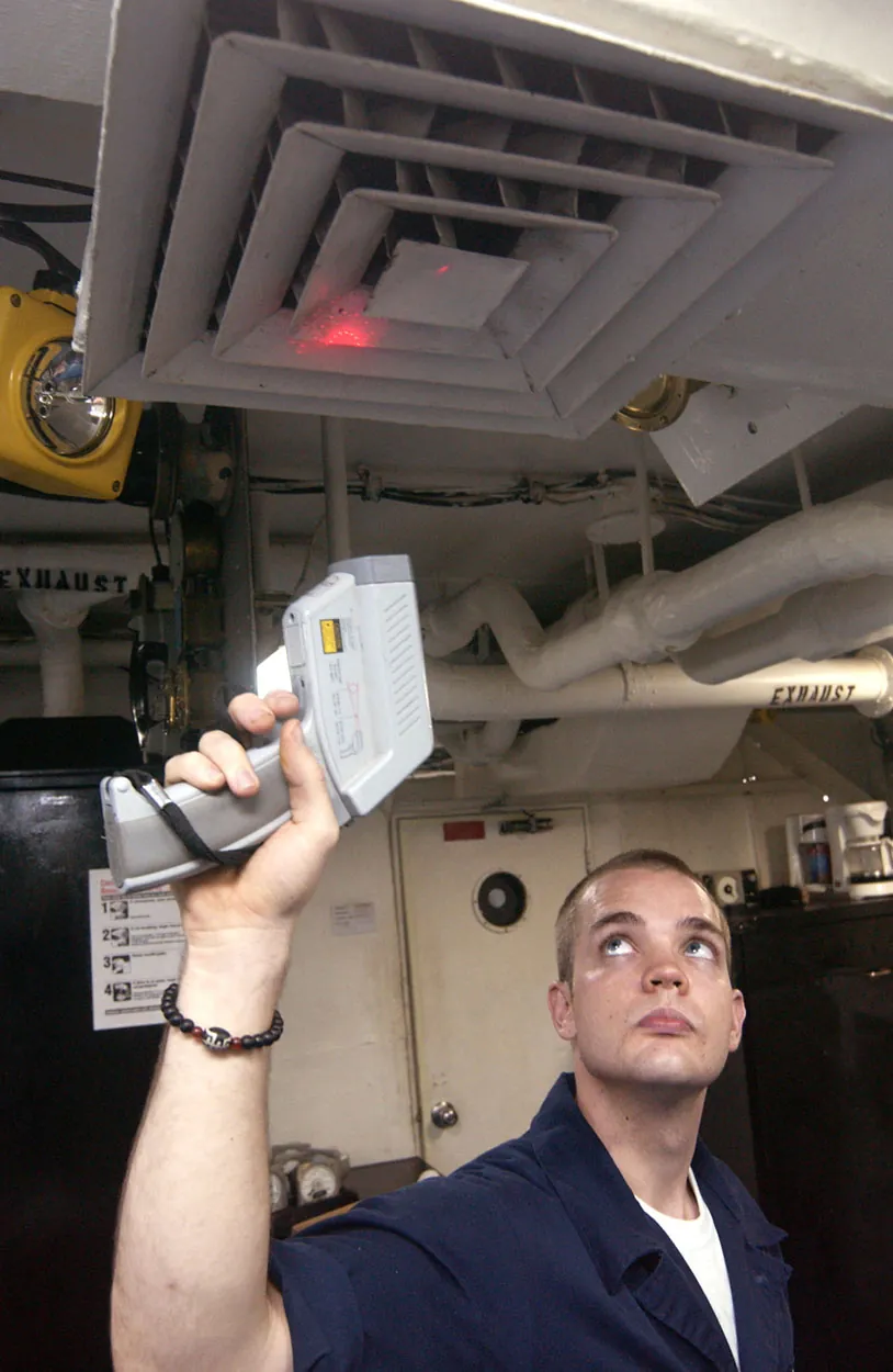 A man holds a device that looks like a gun or a check-out scanner up toward an air vent. A red light emanates from the device and shines on the vent.